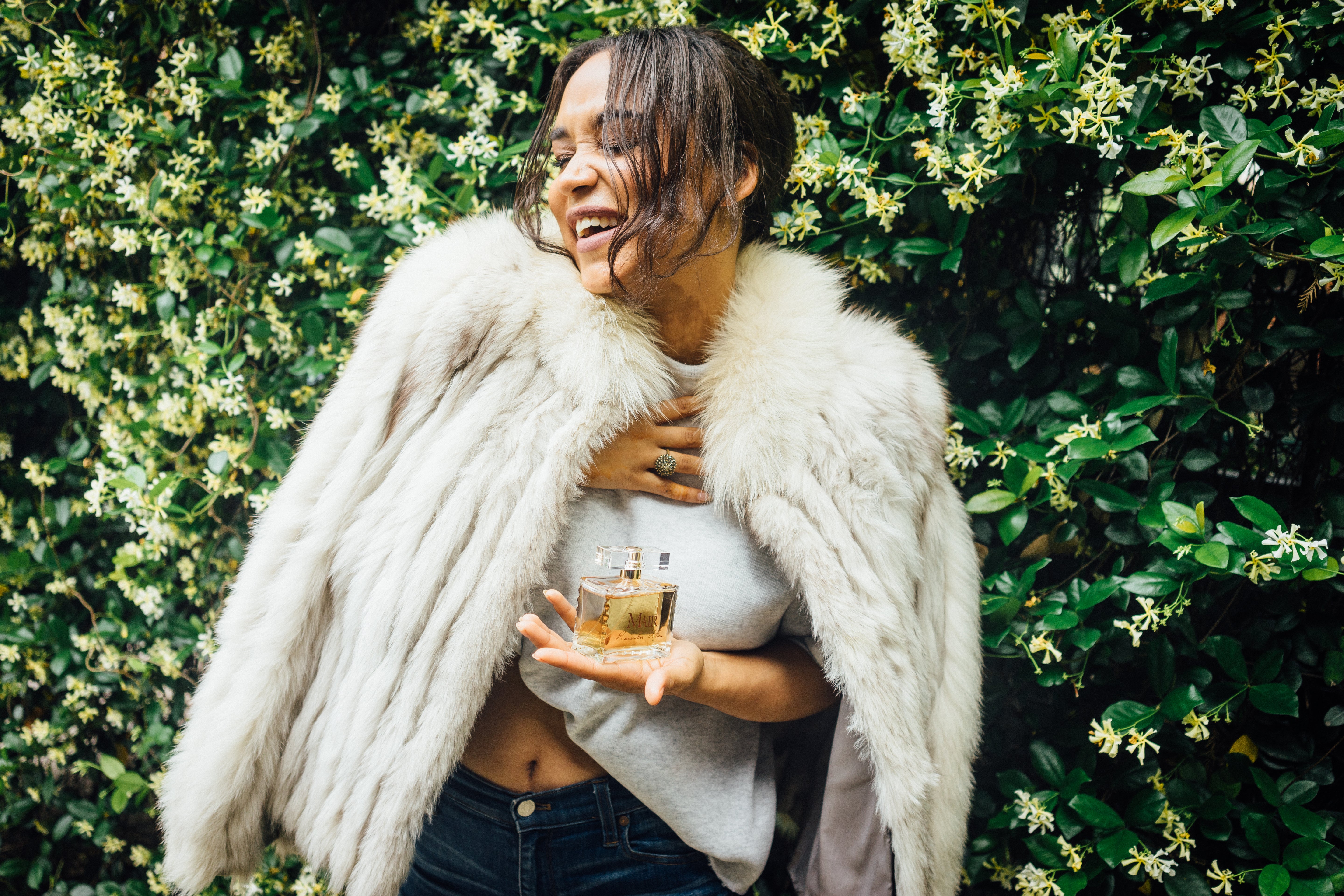 Woman in a white fur coat holding a Mair Perfume bottle against a green leafy background