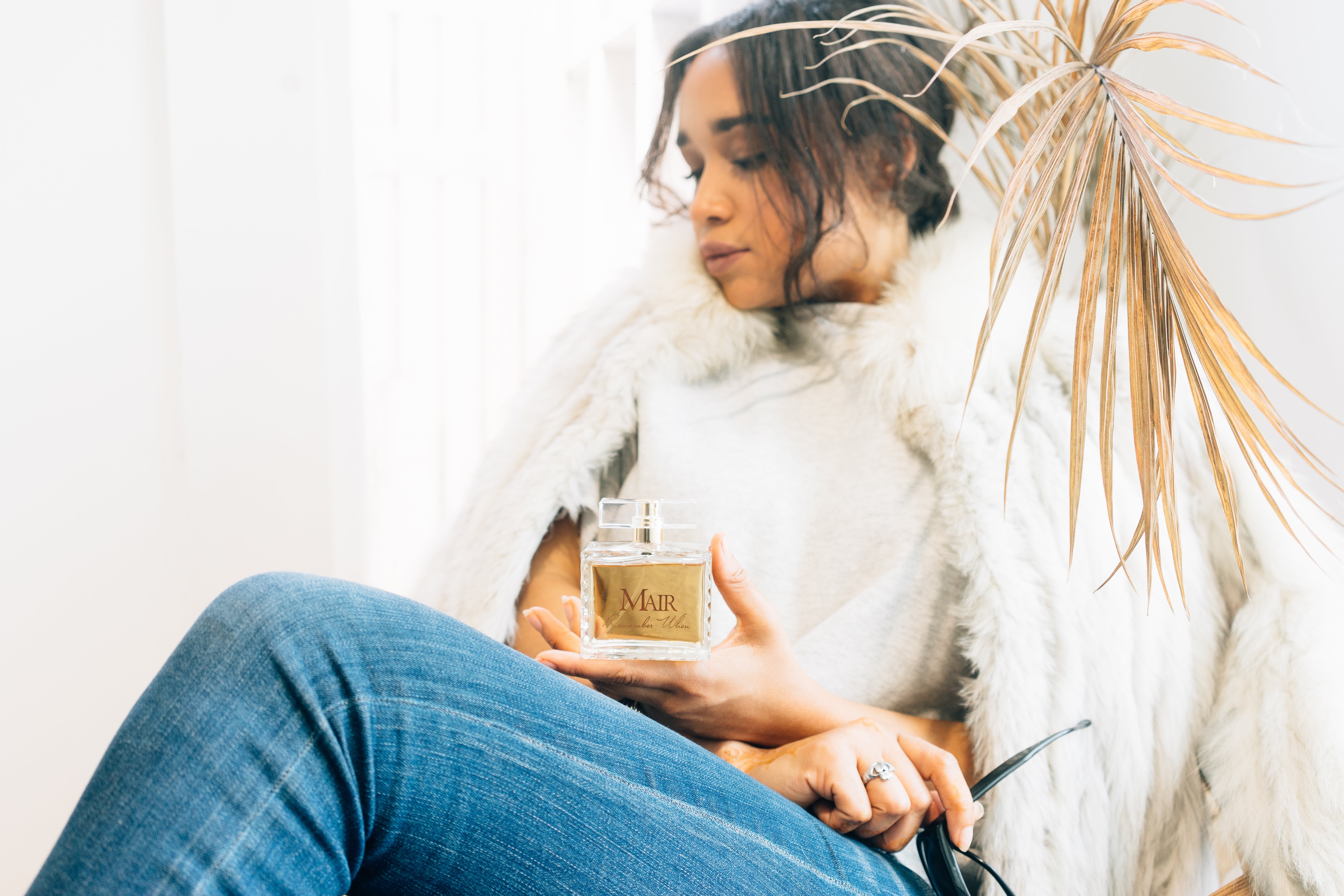 Woman holding a candle labeled 'Mair' in a cozy setting with a white blanket and decorative plant.